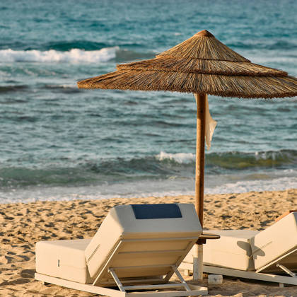 Beach scene with two cushioned sun loungers under a straw umbrella, ocean waves in the background.